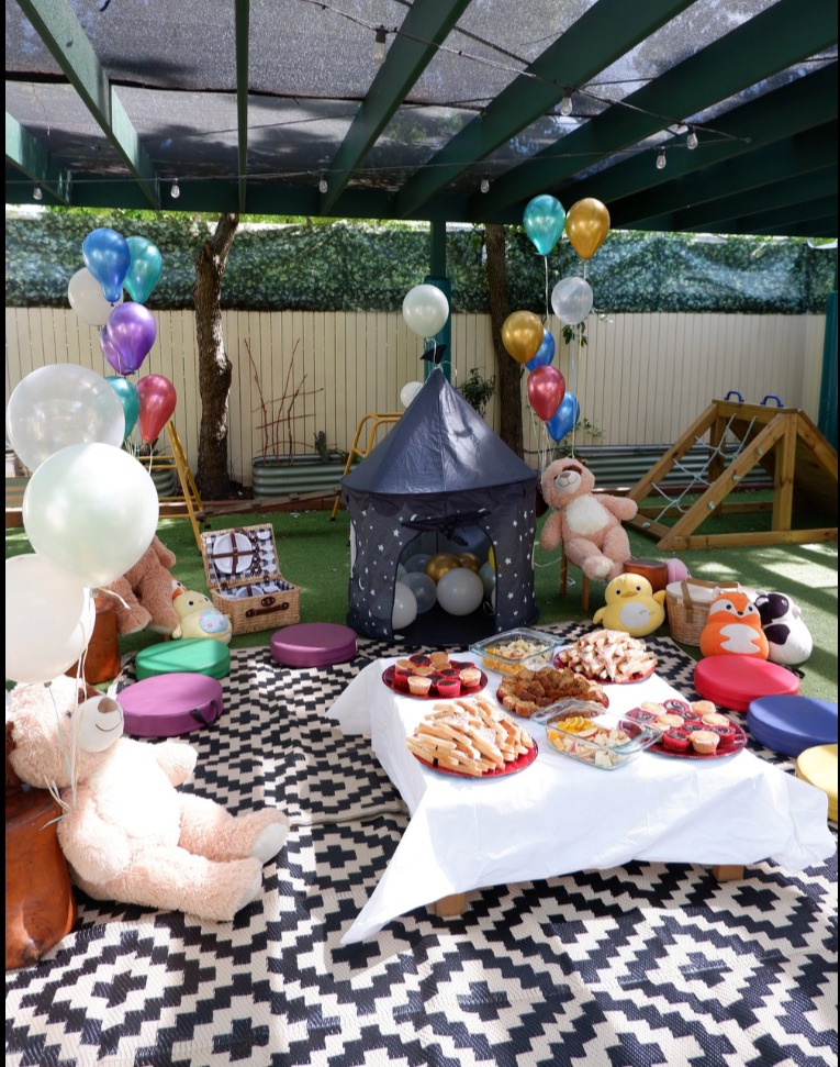 Teddy bears picnic set up in the outdoor play space with balloons, cushions and a shared table of food